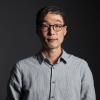 Taeho Kim wearing a grey button-down shirt with glasses sitting for his headshot in front of a black backdrop.